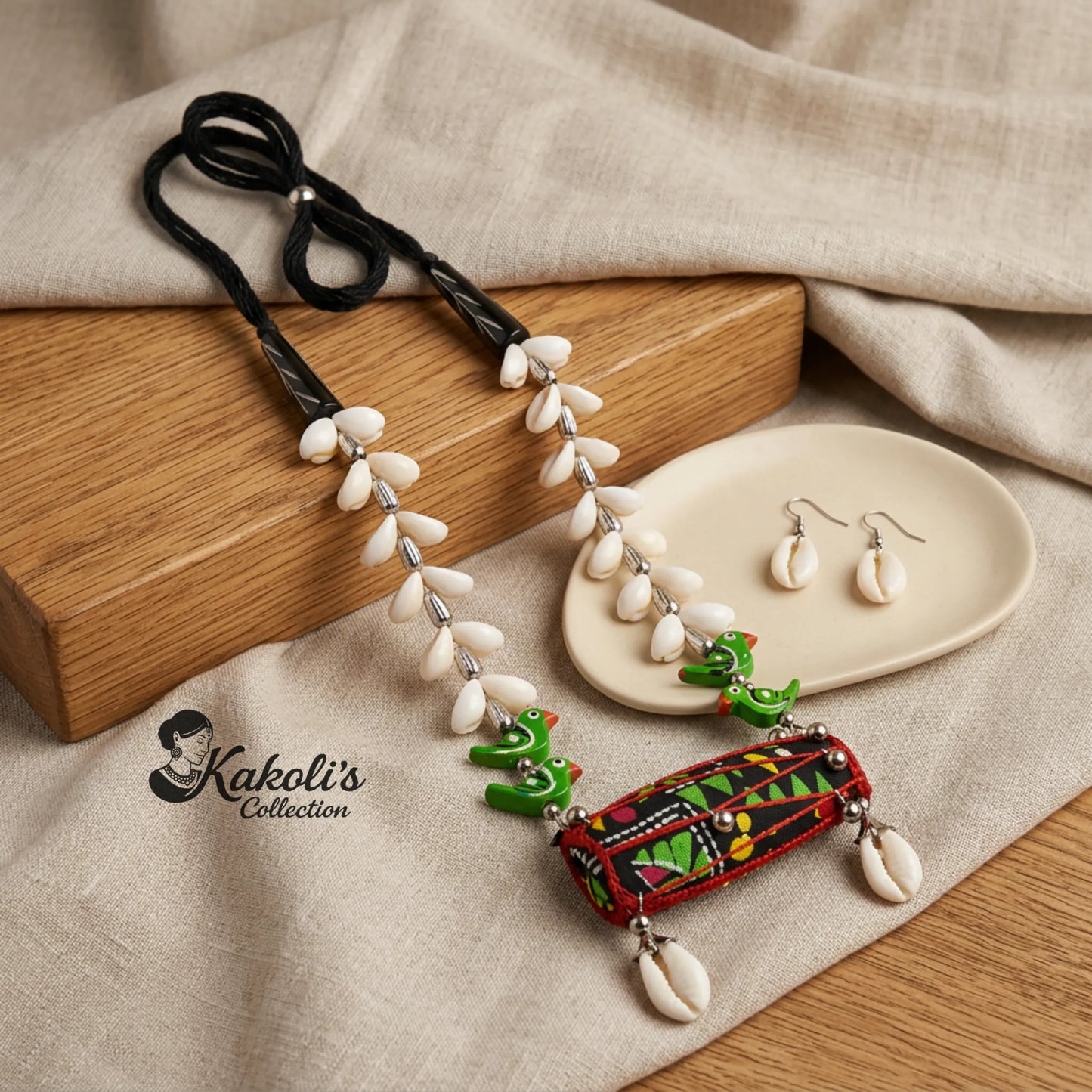 Red and Green Tribal Rhythms Dhol and Cowrie Handmade Fabric Necklace Set is placed  on a textured surface with 'Kakoli's Collection' branding.